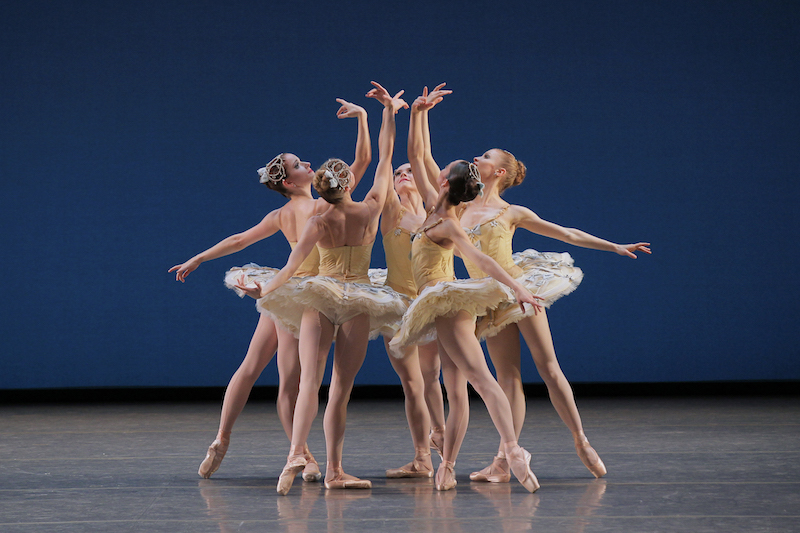Five ballerinas stand into a circle wearing yellow jewel encrusted tutus. They look into the palm of their right raised arm.